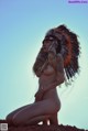 A naked woman wearing an indian headdress sitting on a rock.