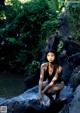 A woman in a bathing suit sitting on a rock by a river.